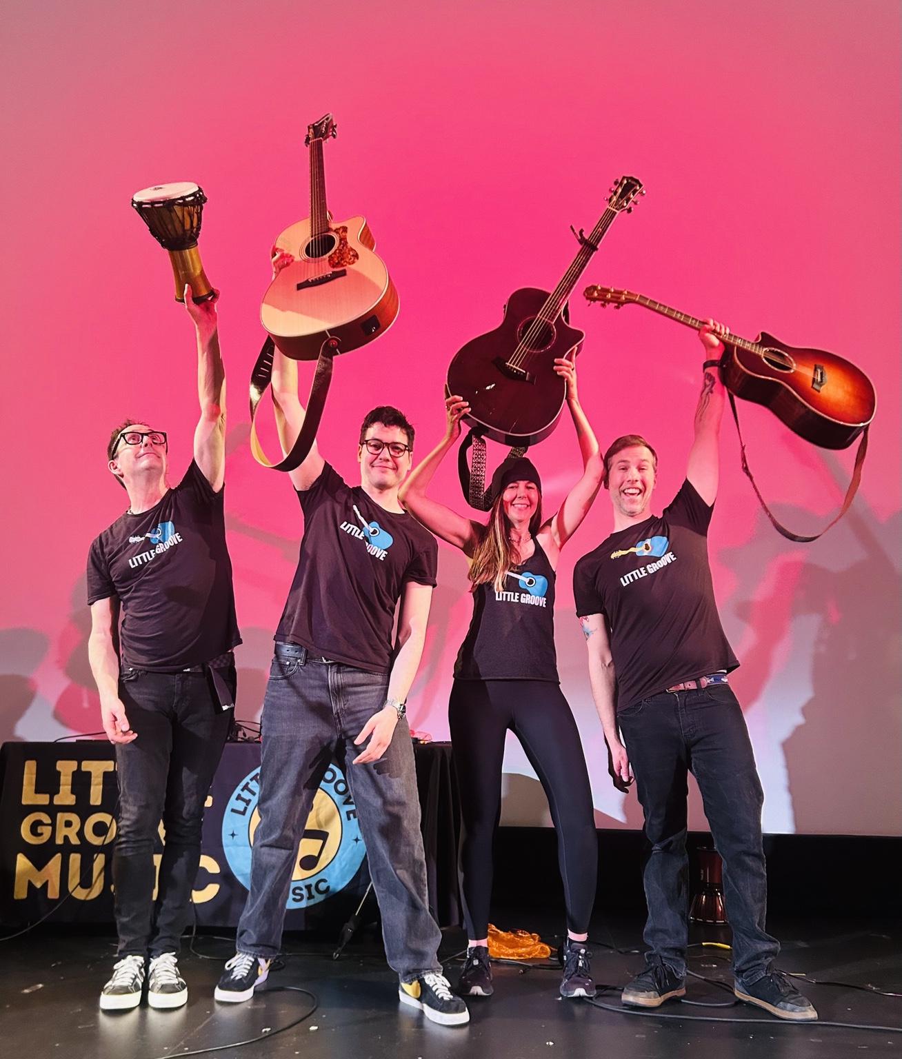 Little Groove musicians onstage holding guitars and a drum high against a pink backdrop